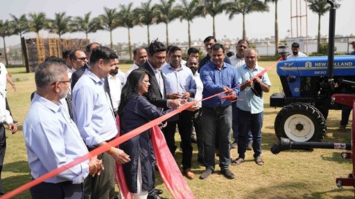 A group of people gathered outdoors on a grassy area with palm trees in the background, participating in a ribbon-cutting ceremony. A woman and a man in the center are cutting a red ribbon with scissors, while others stand around them watching. A blue New Holland tractor is visible on the right side of the image. The event appears to be a formal or celebratory occasion.