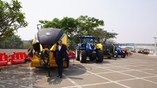 A man in a dark suit stands next to large yellow and black agricultural machinery and blue tractors parked in a row on a paved outdoor area with trees and red barriers in the background under a clear sky.