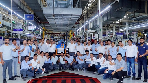 A large group of factory workers and engineers posing together inside a manufacturing facility, all giving thumbs-up gestures. They are standing and squatting around a blue tractor assembly line area, with industrial equipment and machinery visible in the background. The environment is well-lit with overhead lights and organized workstations.