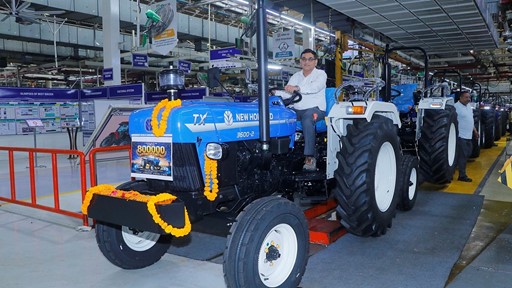 A man sitting on a blue New Holland TX 3600-2 tractor decorated with orange marigold garlands inside a factory assembly line. The tractor has a sign indicating "800000" units produced. Several similar tractors are lined up behind it, and factory workers are visible in the background. The setting is a well-lit industrial environment with various signs and equipment.