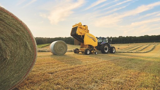A blue tractor with a yellow hay baler attachment is working in a large open field during sunset, creating round hay bales. One large hay bale is in the foreground on the left, with more hay bales scattered across the field and a line of trees in the background under a partly cloudy sky.