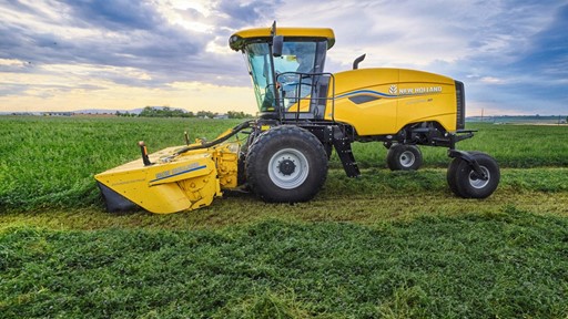A yellow New Holland agricultural harvester machine is parked on a green field under a partly cloudy sky during daylight, with freshly cut crops visible on the ground.