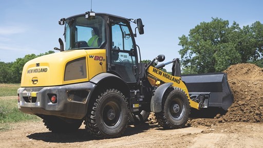 Yellow New Holland W80D wheel loader with a front bucket filled with dirt, parked on a construction site with trees and a clear blue sky in the background.
