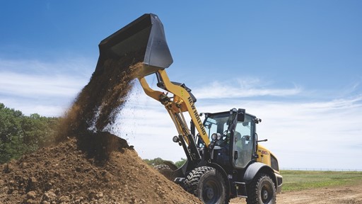 A yellow New Holland wheel loader is dumping a load of dirt onto a large pile at a construction site under a clear blue sky.
