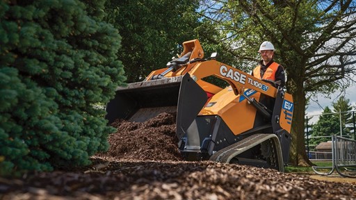 A worker wearing a white hard hat and orange safety vest operates a yellow CASE TL100EV electric mini track loader spreading mulch around a landscaped area with trees and greenery in the background