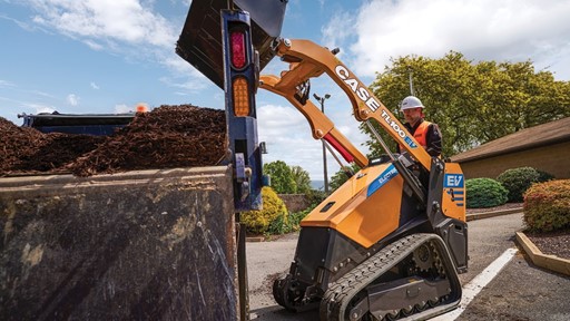 A worker wearing a white hard hat and orange safety vest operates a yellow CASE TL100EV electric mini track loader dumping mulch into a large container on a sunny day with blue sky and scattered clouds