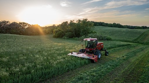 A red agricultural mower cutting grass in a large green field during sunset, with trees and rolling hills in the background under a partly cloudy sky.