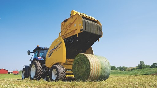 A blue tractor attached to a yellow New Holland round baler machine is operating in a green field under a clear blue sky, producing a large round bale of hay. In the background, there are some trees and a red barn.