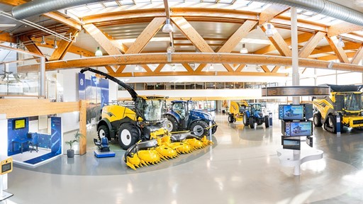 Interno di un moderno showroom agricolo con macchinari agricoli di grandi dimensioni, tra cui trattori blu, mietitrebbie gialle e attrezzature per la raccolta, illuminato da ampie finestre e travi in legno a vista.