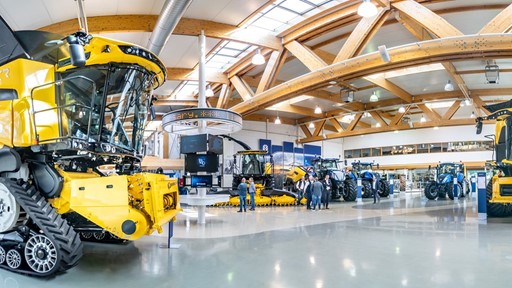 Interno di un grande showroom con macchinari agricoli moderni di colore giallo e blu, tra cui mietitrebbie e trattori, con alcune persone che osservano e discutono vicino ai veicoli, struttura con travi in legno e ampie finestre che lasciano entrare molta luce naturale.