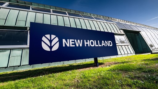Insegna blu con il logo e la scritta "NEW HOLLAND" davanti a un edificio moderno con pareti in metallo verde e finestre, su un prato verde sotto un cielo sereno.