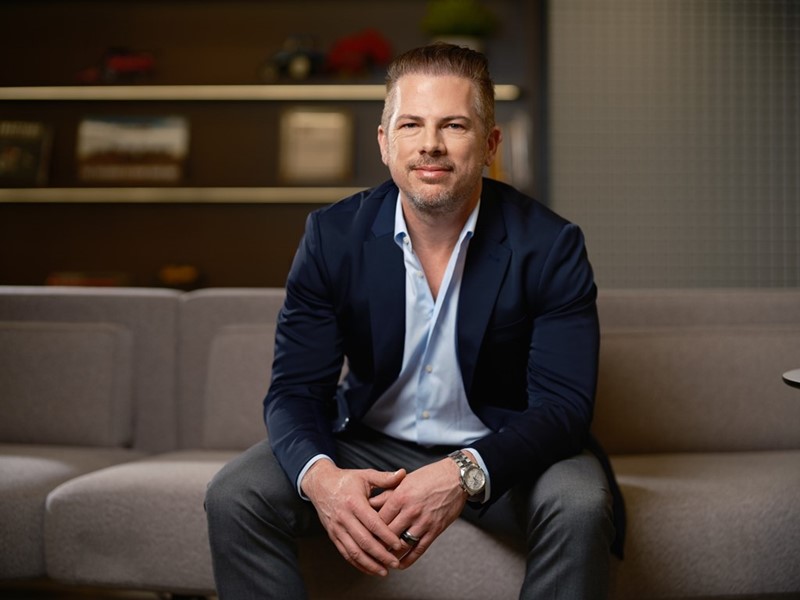 A professional man stands confidently in a modern office environment. He is wearing a dark blazer over a light blue shirt, with a slight smile on his face. The background features a blurred office space with plants and soft lighting, creating a welcoming atmosphere. The man is leaning against a wooden table, exuding a sense of professionalism and approachability.