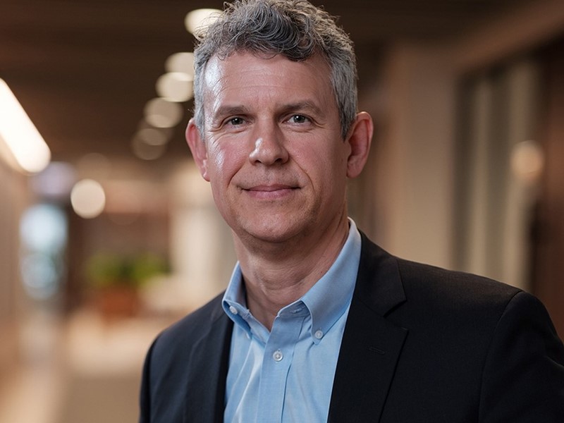 Portrait of a middle-aged man with gray curly hair wearing a light blue button-up shirt and a black blazer, standing in a softly lit indoor hallway with blurred background.