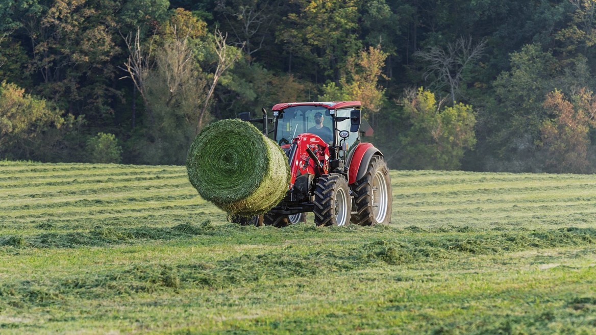 A red tractor pulling a large round hay bale across a lush green field, surrounded by trees and a clear sky. The scene captures the essence of agricultural work and rural life.