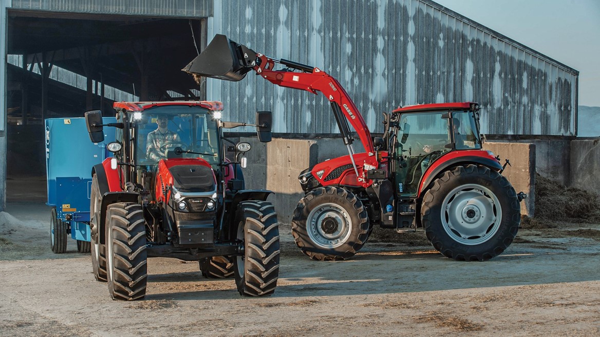 Two red tractors are parked in a farmyard. The tractor on the left has a front loader attachment and a driver inside, while the tractor on the right is equipped with a rear implement. In the background, a large barn with a metal roof is visible, and the ground is dusty, indicating an agricultural setting.