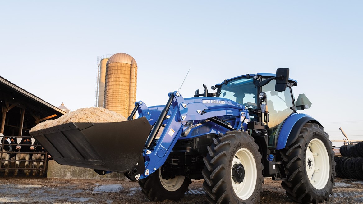 A blue New Holland tractor with a front loader is positioned on a farm, carrying a large bucket of material. In the background, a silos and a barn are visible under a clear sky, indicating a rural agricultural setting. The tractor's tires are large and rugged, suitable for farm work.
