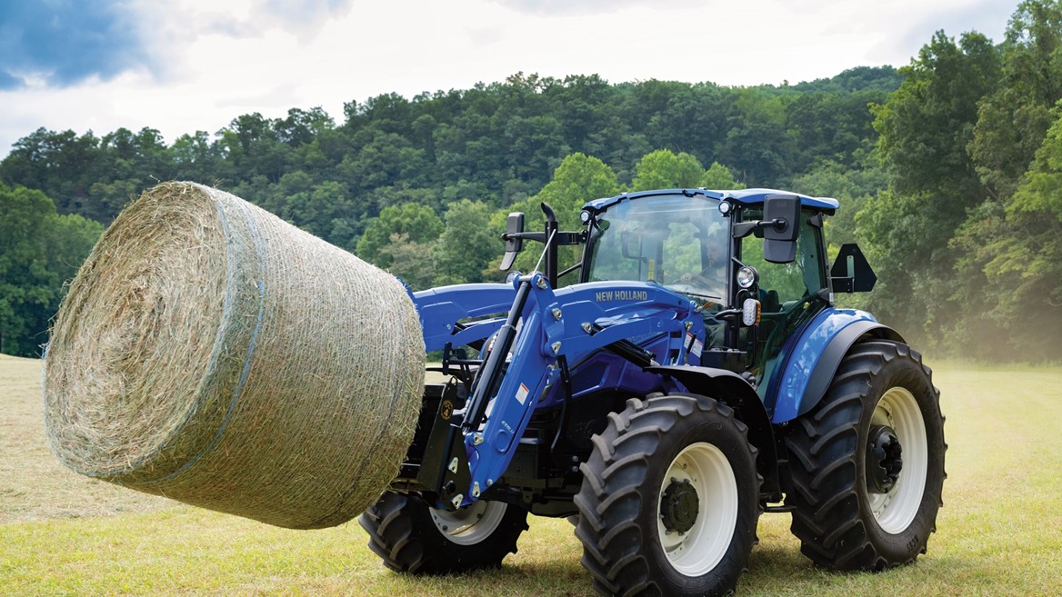 A blue New Holland tractor is lifting a large round hay bale in a grassy field. The background features lush green trees and a cloudy sky, indicating a rural farming setting. The tractor's front loader is engaged, showcasing its capability for handling heavy loads.