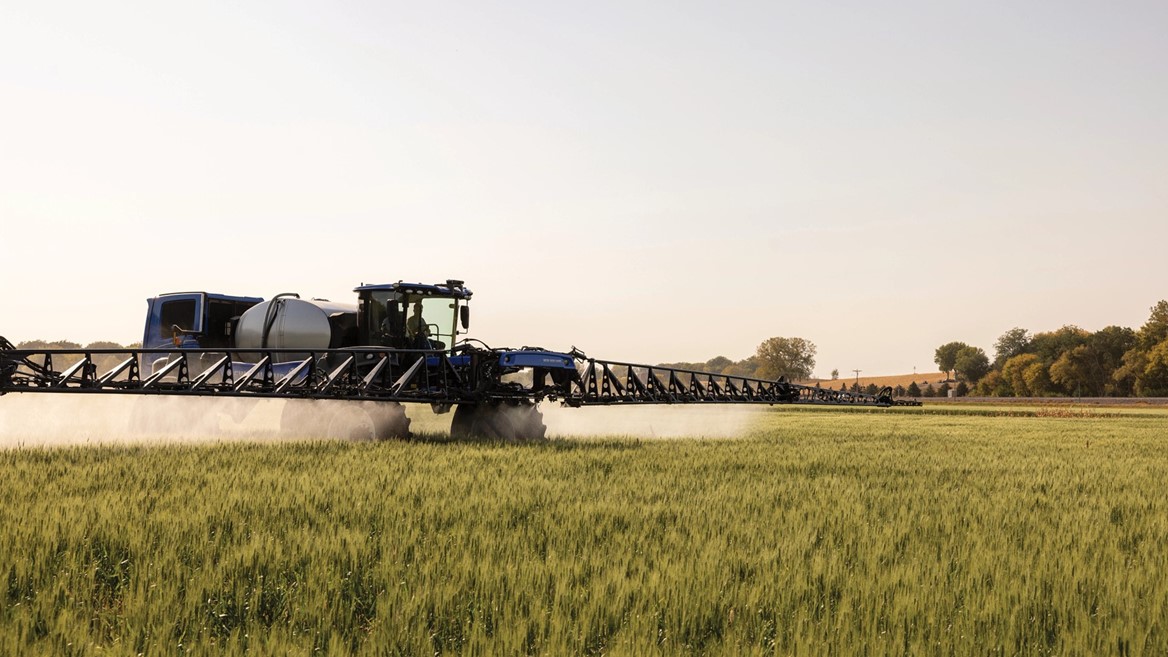 A blue agricultural tractor equipped with a large sprayer is working in a green field during a clear day. The tractor is spraying a mist over the crops, with trees and a clear sky in the background.