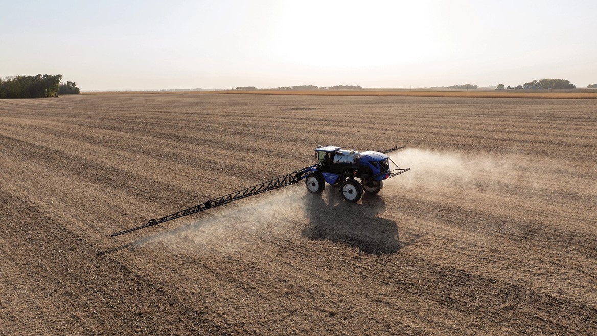 A blue tractor equipped with a sprayer is working in a vast, empty agricultural field. Dust is being kicked up as the tractor moves across the dry, brown soil under a clear sky. The landscape is flat and stretches into the distance, showcasing the agricultural setting.