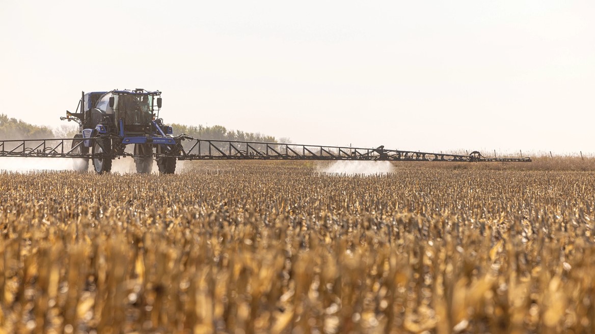 A large agricultural sprayer tractor applying chemicals to a field of dry, golden crops under a clear sky. The tractor is positioned in the distance, with a long spray arm extending outward, creating a mist as it moves through the harvested field.