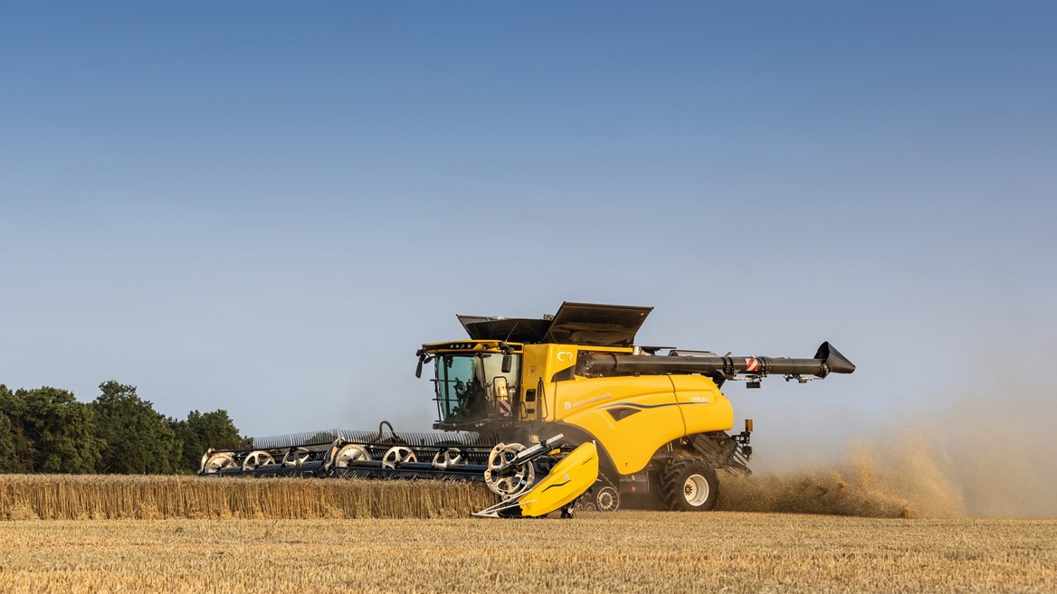 A yellow combine harvester working in a field during harvest season, with dust rising as it cuts through golden wheat under a clear blue sky.