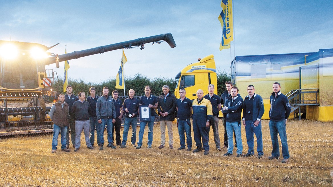 A group of 15 people stands in a field during twilight, with a large agricultural machine and a truck in the background. The individuals are dressed in casual work attire and are smiling, holding a plaque. Yellow flags with a logo are visible, indicating a celebration or event related to farming or agriculture. The setting appears to be a harvest or agricultural demonstration.