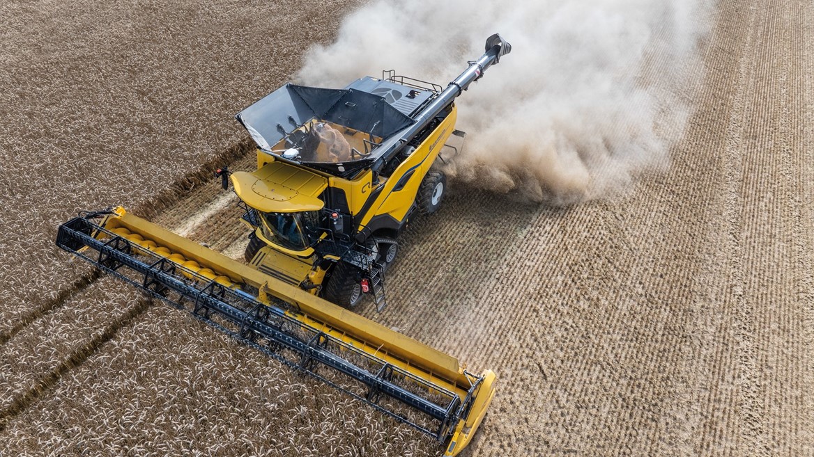 Aerial view of a yellow combine harvester working in a field, with dust and debris being kicked up as it cuts through rows of crops. The machine is equipped with a wide header, efficiently harvesting the agricultural produce. The surrounding field shows neatly arranged crop rows, highlighting the agricultural landscape.