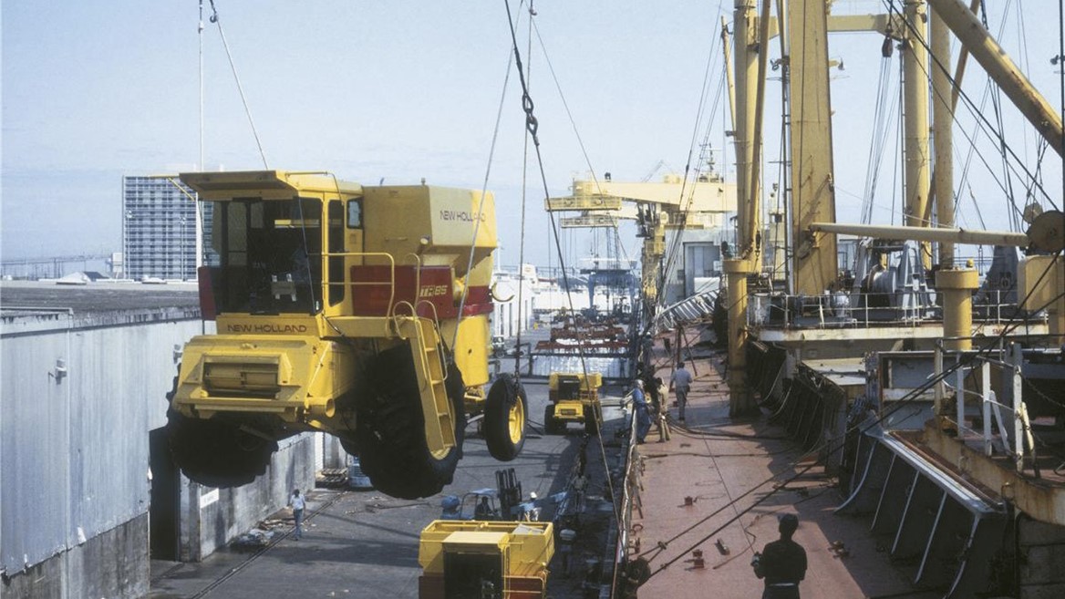 A yellow agricultural harvester being lifted by a crane at a port, with cargo ships and workers in the background. The scene depicts industrial activity, showcasing the transportation of heavy machinery.