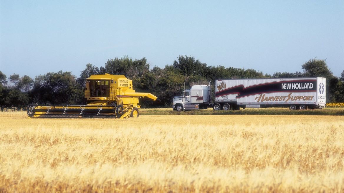 A yellow combine harvester is working in a golden wheat field, with a large truck bearing the "New Holland Harvest Support" logo parked nearby. The background features trees and a clear blue sky, emphasizing a rural agricultural setting.