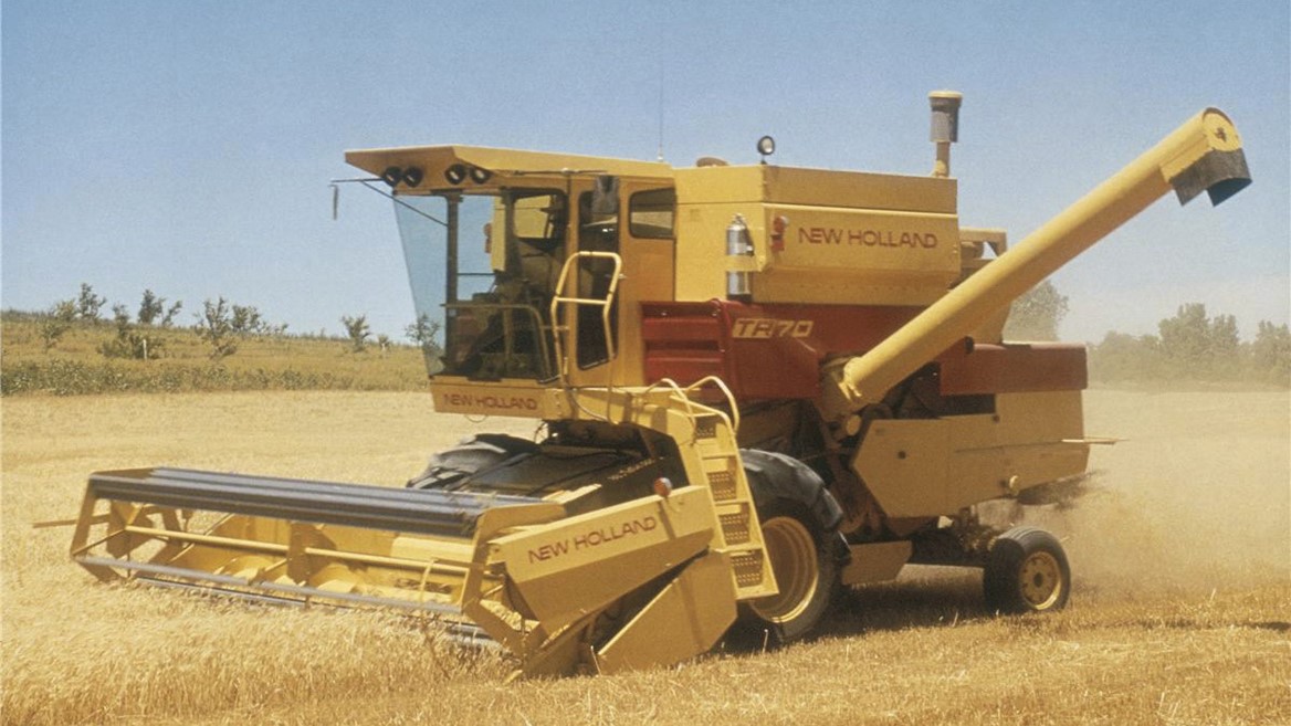 A yellow New Holland combine harvester working in a golden wheat field under a clear blue sky. The machine is actively harvesting crops, with dust rising around it, showcasing agricultural machinery in action.