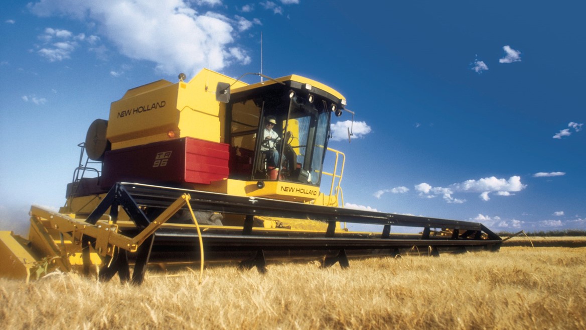 A New Holland combine harvester working in a golden wheat field under a blue sky with scattered clouds. The machine is prominently featured, showcasing its yellow and red colors as it cuts through the tall wheat.