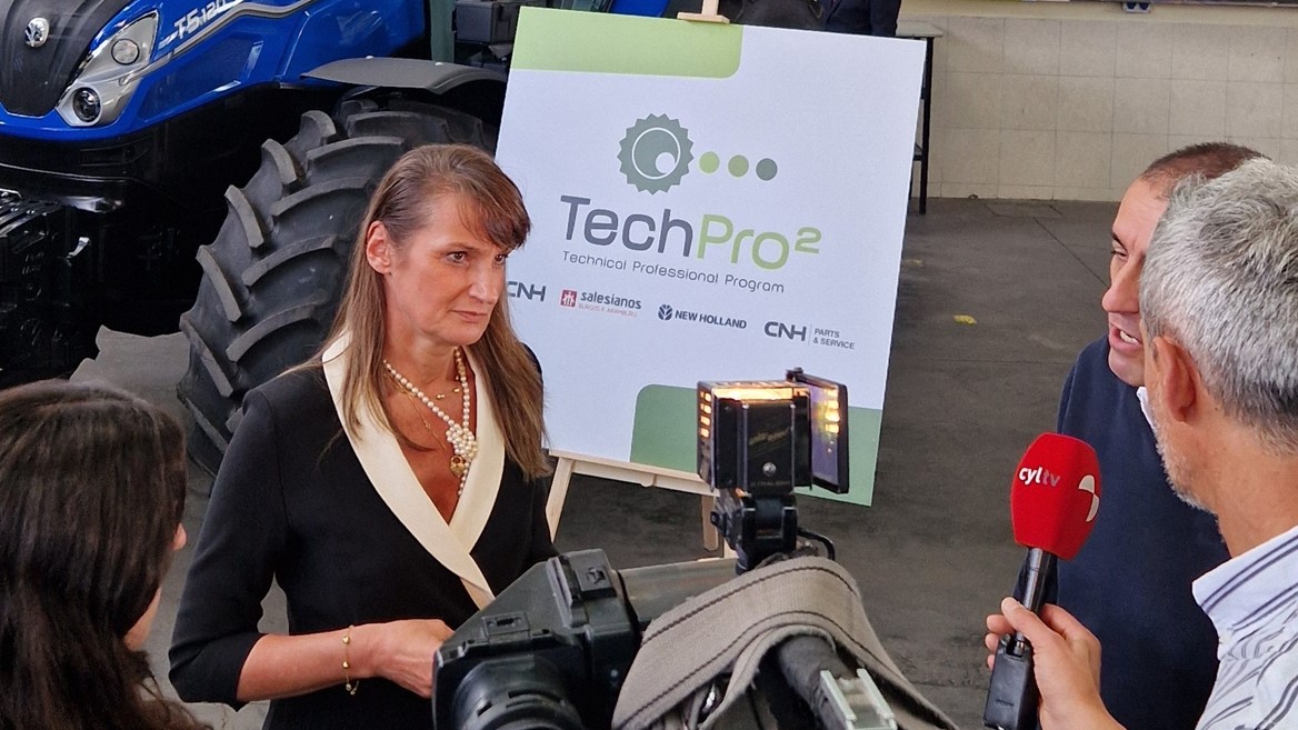 A woman is being interviewed by a news crew inside a workshop or showroom with a blue New Holland tractor in the background. Behind her is a large sign displaying the "TechPro2" logo and the text "Technical Professional Program." The setting includes posters of people working on machinery, emphasizing a technical or agricultural theme. The interviewers hold microphones and a professional video camera is recording the scene.