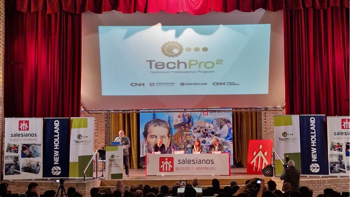A large audience seated in an auditorium facing a stage with red curtains. On the stage, a speaker stands at a podium with a microphone, presenting in front of a large screen displaying the "TechPro² Technical Training Program" logo. Banners for Salesianos, New Holland, and CNH Industrial flank the stage. The event appears to be a formal presentation or training session.