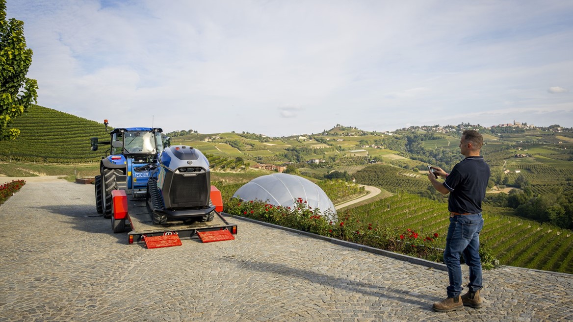 A man operating a remote control stands on a cobblestone path overlooking a scenic vineyard landscape with rolling hills. In front of him, a blue tractor is carrying a modern robotic agricultural machine on a trailer. The vineyard rows stretch across the hills under a partly cloudy sky, with a white dome structure visible among the greenery.