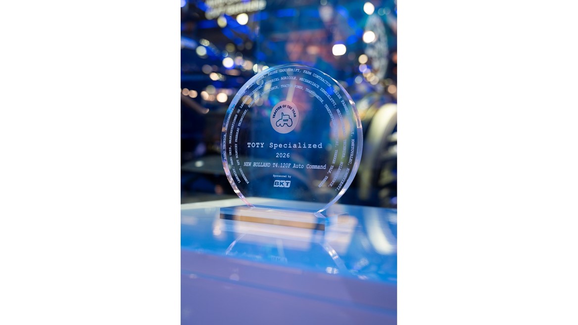 A clear circular glass award trophy with the inscription "TOTY Specialized 2026 NEW HOLLAND T4.120F Auto Command" and the logo "Tractor of the Year" featuring a tractor icon. The award is sponsored by BKT and is displayed on a reflective surface with a blurred background of blue and purple lights.