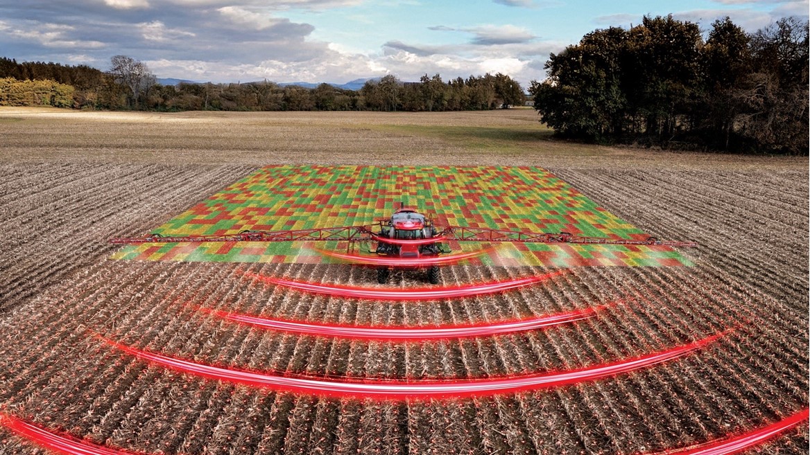 A red agricultural tractor equipped with advanced technology is spraying a field, with a digital overlay showing a colorful grid pattern on the crops and red curved lines representing sensor or radar signals, illustrating precision farming and smart agriculture techniques in a rural landscape under a partly cloudy sky.