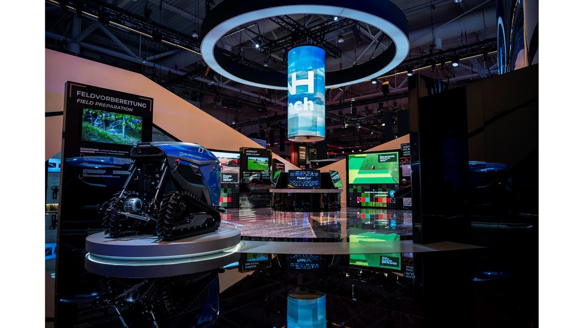 Exhibition booth showcasing advanced agricultural technology with a blue autonomous robotic vehicle on a circular platform, multiple digital screens displaying field preparation and seed planting information, and a large cylindrical hanging digital display with the word "Tech" in a modern indoor event space.