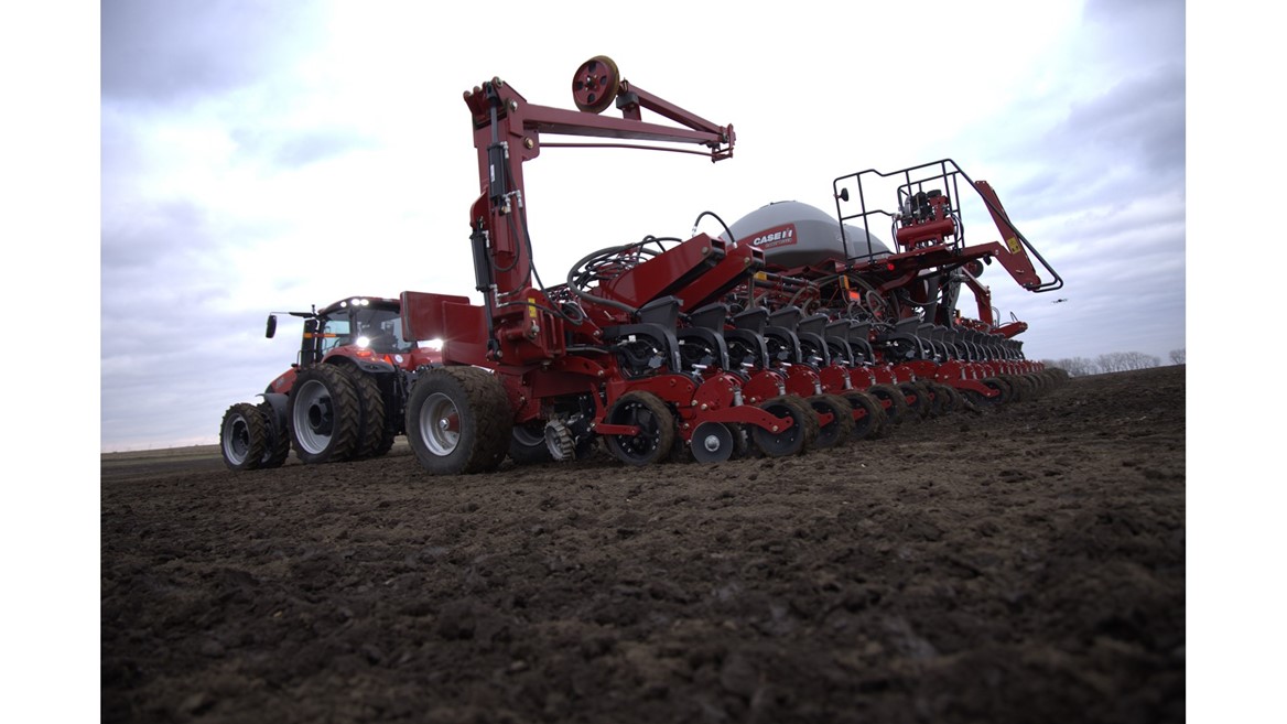 A large red agricultural tractor attached to a multi-row planter machine working on a freshly tilled field under a cloudy sky, preparing the soil for planting crops.