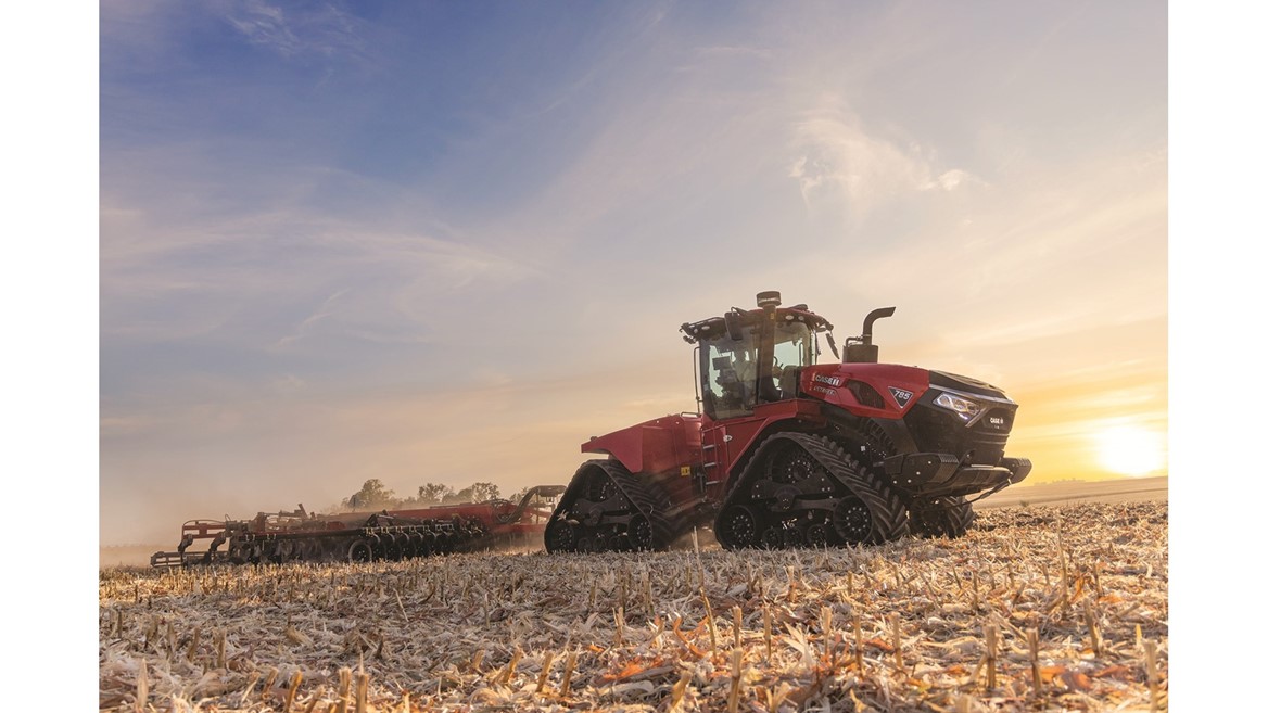 A large red Case IH tractor with rubber tracks is working in a harvested field during sunset, pulling farming equipment behind it under a partly cloudy sky.