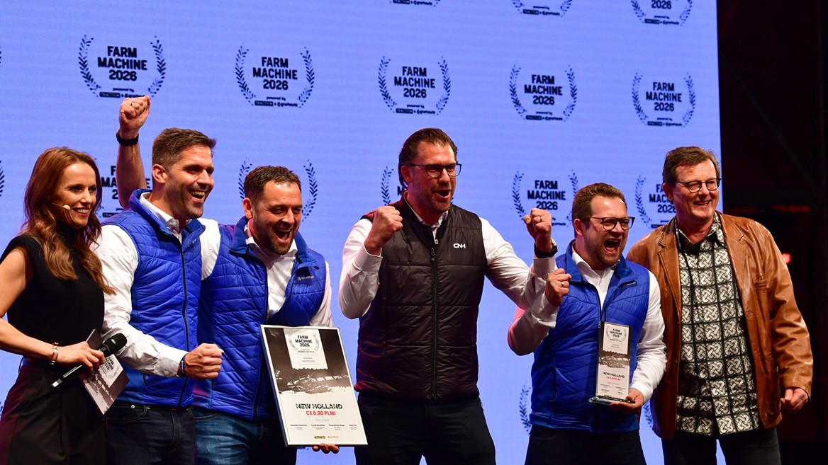 A group of six people standing on stage celebrating an award. Four men in blue and black vests are holding plaques and certificates, smiling and raising their fists in excitement. A woman on the left holds a microphone and a man on the right wears a brown leather jacket and patterned shirt. The background displays multiple "Farm Machine 2026" logos.