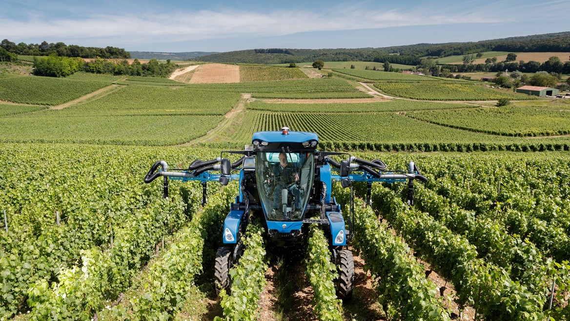 A blue agricultural machine is operating in a vineyard with rows of green grapevines under a clear blue sky, surrounded by rolling hills and farmland in the background.