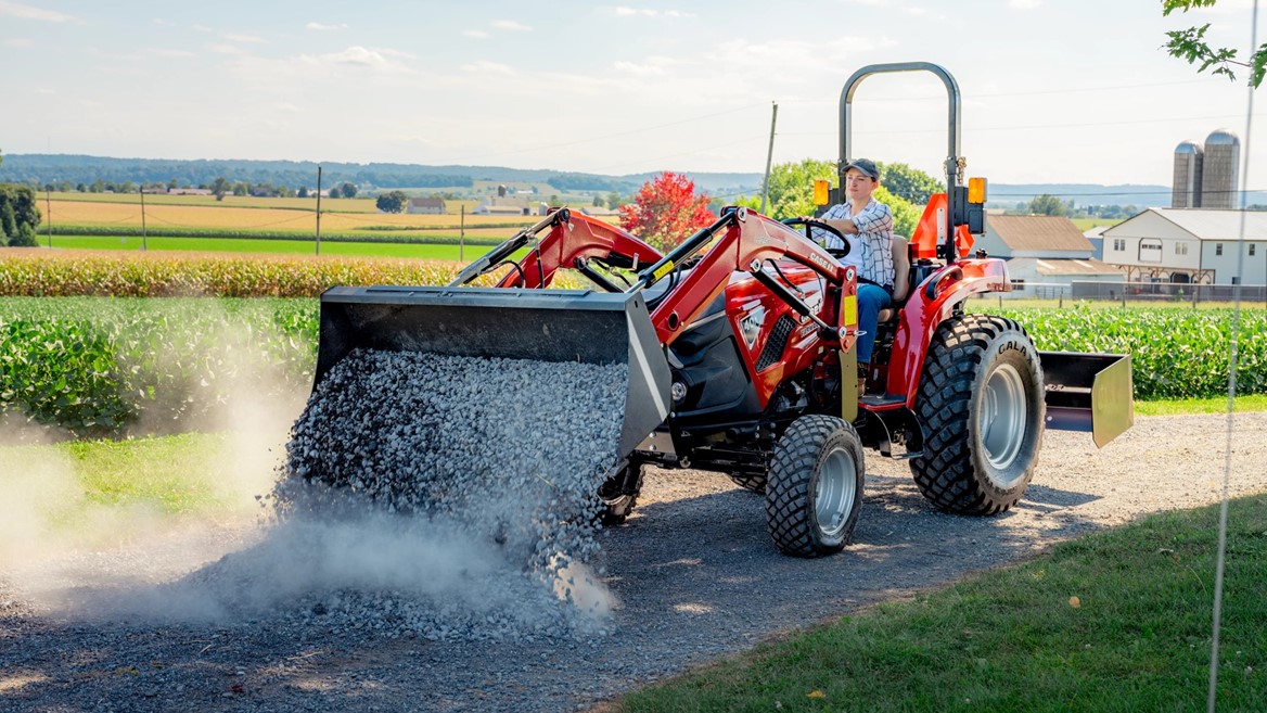 A person operating a red tractor with a front loader, spreading gravel on a rural dirt road. The background features green fields, a farmhouse, silos, and a clear blue sky with scattered clouds.