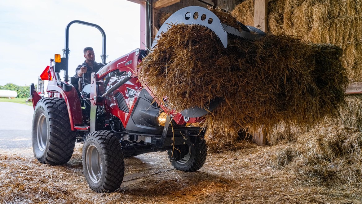 A person operating a red tractor inside a barn, using a front loader attachment to lift a large bale of hay. The barn is filled with stacked hay bales and straw scattered on the ground.