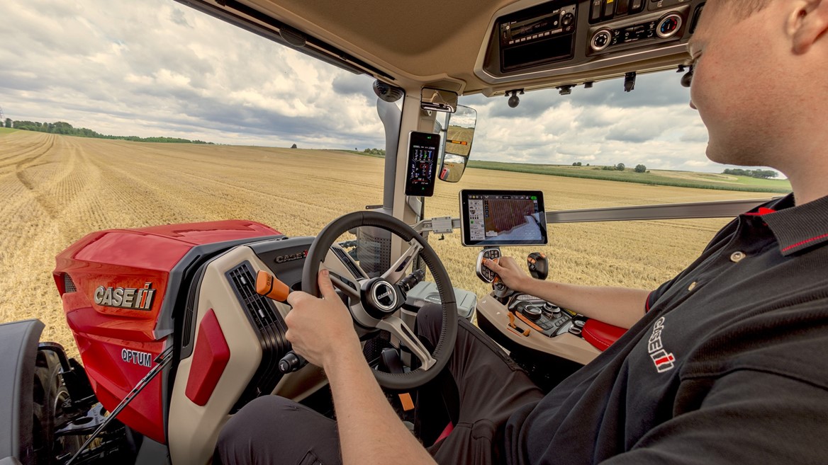 Interior view of a modern Case IH Optum tractor cabin showing a person operating the steering wheel and control panel, with a large golden wheat field visible through the windows under a cloudy sky.