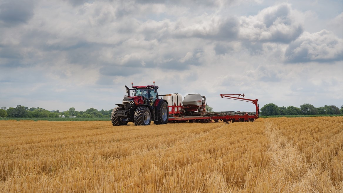 A red tractor with large tires is working in a golden wheat field under a cloudy sky, pulling farming equipment used for planting or fertilizing. Trees and greenery are visible in the distant background.