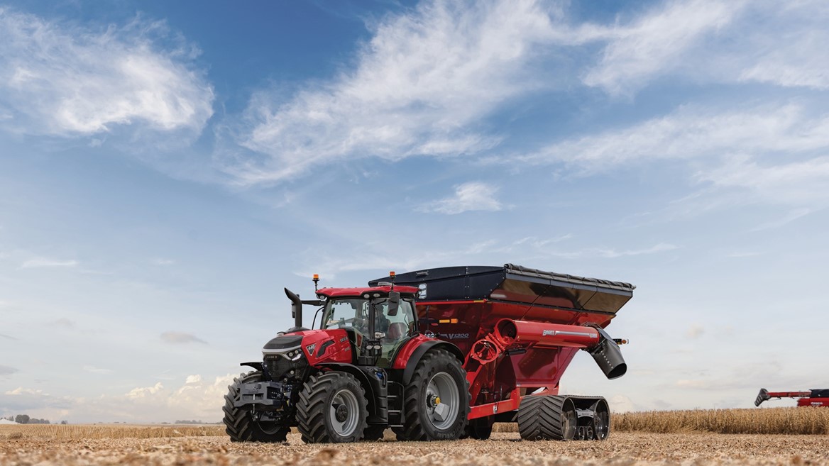 A red tractor attached to a large red grain cart with black tracks is parked in a harvested wheat field under a partly cloudy blue sky.