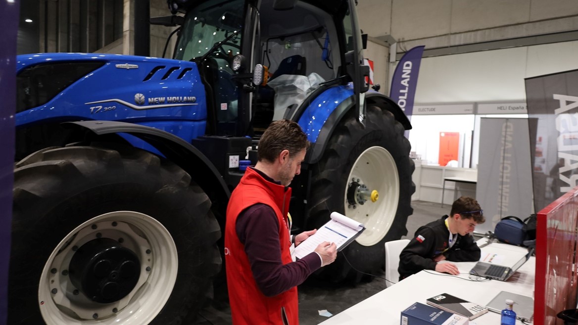 A large blue New Holland T7.300 tractor is displayed indoors at an exhibition or trade show. In the foreground, a man wearing a red vest is holding and writing on a clipboard, while another person is seated at a white table working on a laptop. Various banners and booths are visible in the background.