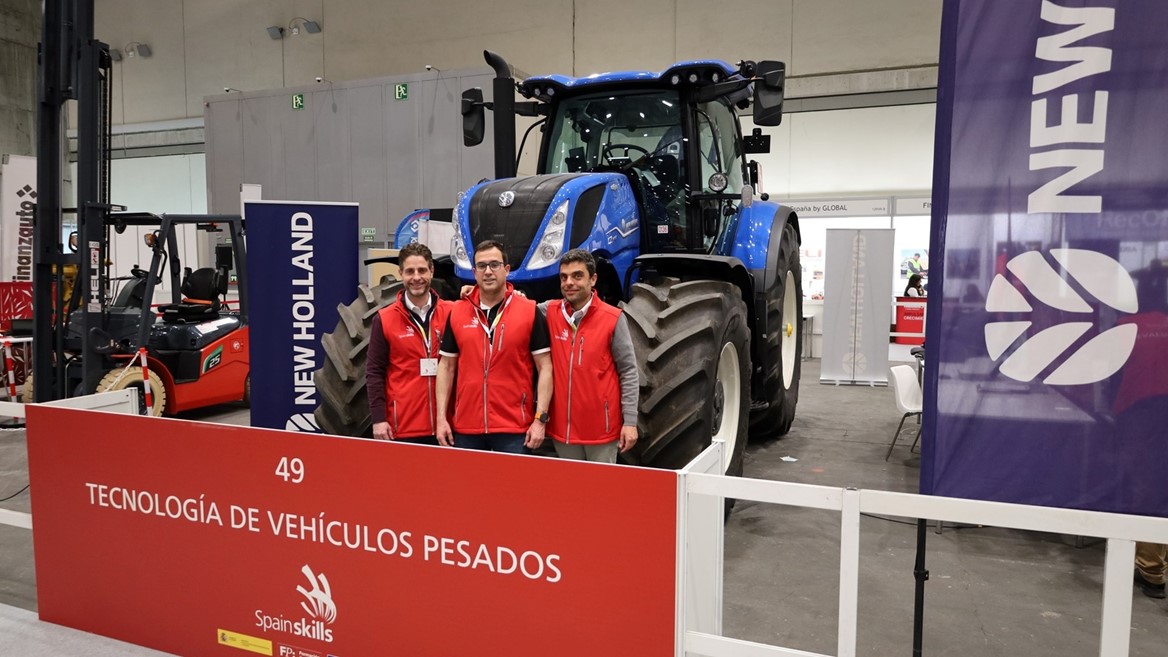 Three men wearing red vests standing in front of a large blue New Holland tractor at an indoor exhibition booth labeled "Tecnología de Vehículos Pesados" with SpainSkills branding.