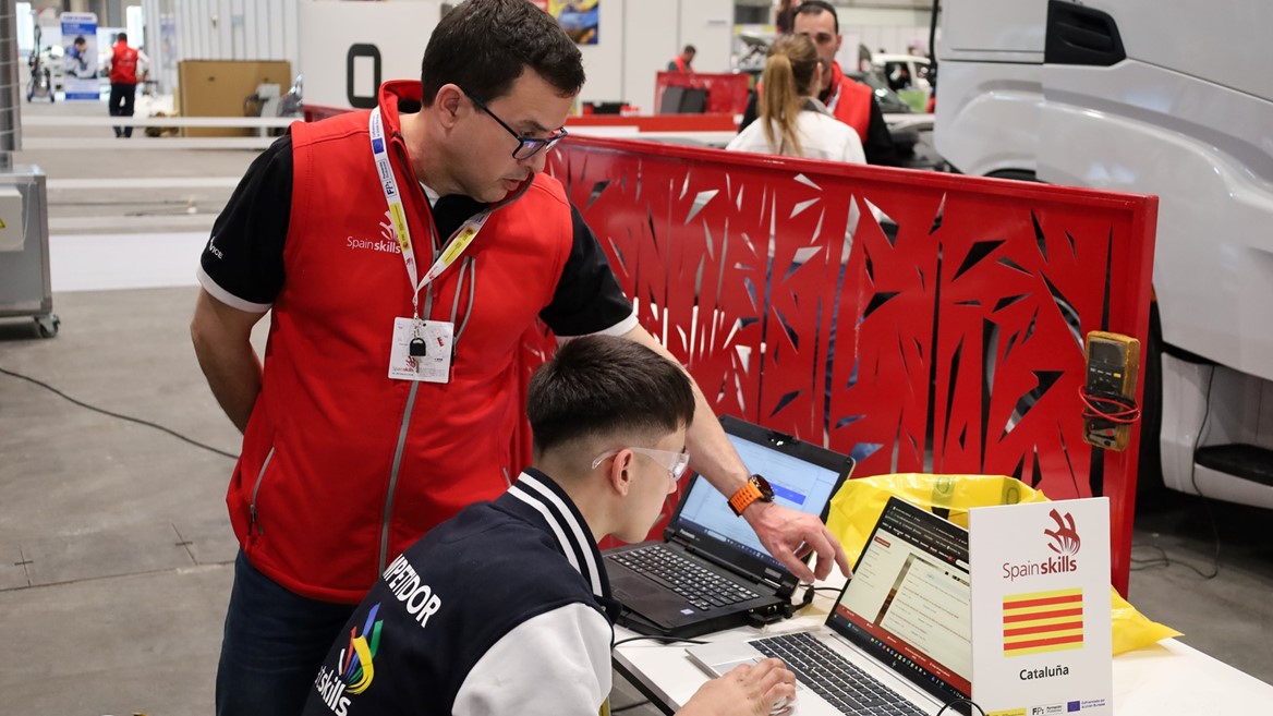 Two men working together on laptops at a SpainSkills event in an indoor workshop setting, with one man standing and pointing at the laptop screen while the other sits and types. A sign on the table displays the SpainSkills logo and the word "Cataluña" with the Catalonia flag. The background shows other participants and equipment in a spacious, well-lit area.