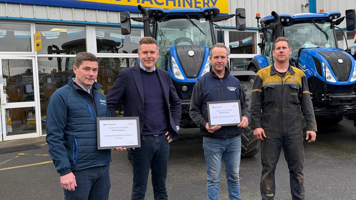 Four men standing in front of two blue tractors outside a building with a yellow and blue "M&S Machinery" sign. Two of the men are holding framed certificates, and all are dressed in casual or work attire. The scene appears to be an award presentation or recognition event.